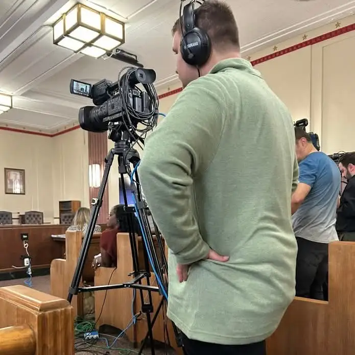 A camera on a tripod covering a public meeting from the side of the room