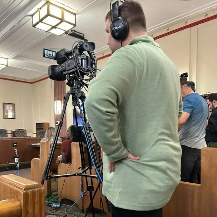 A camera on a tripod covering a public meeting from the side of the room
