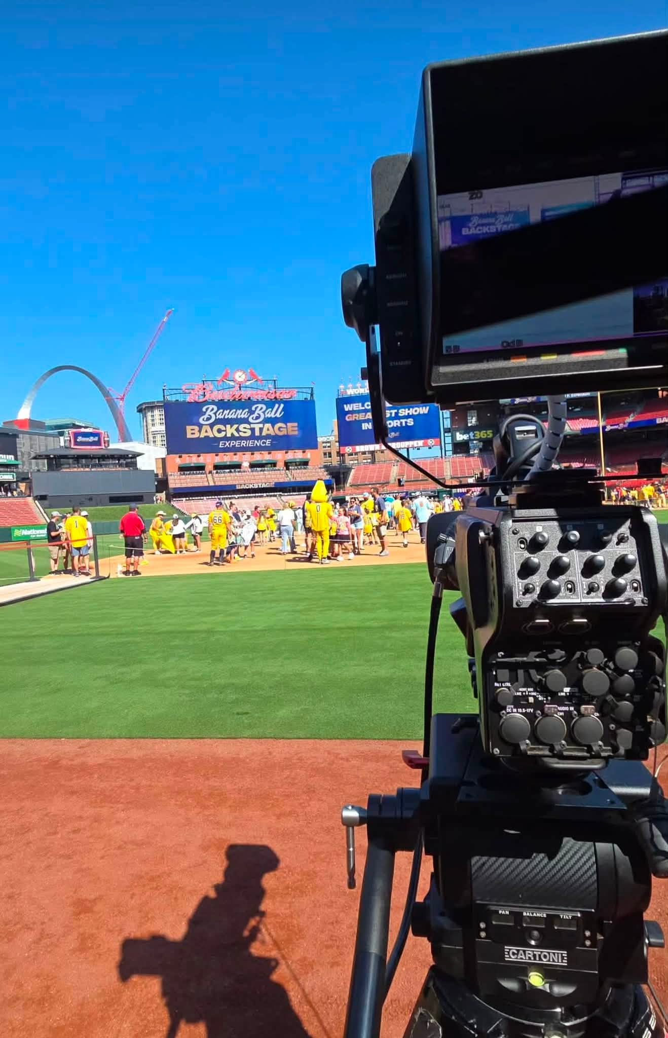 Broadcast camera on a field-level tripod at Busch Stadium during the Banana Ball Backstage Experience, with the Gateway Arch in the background